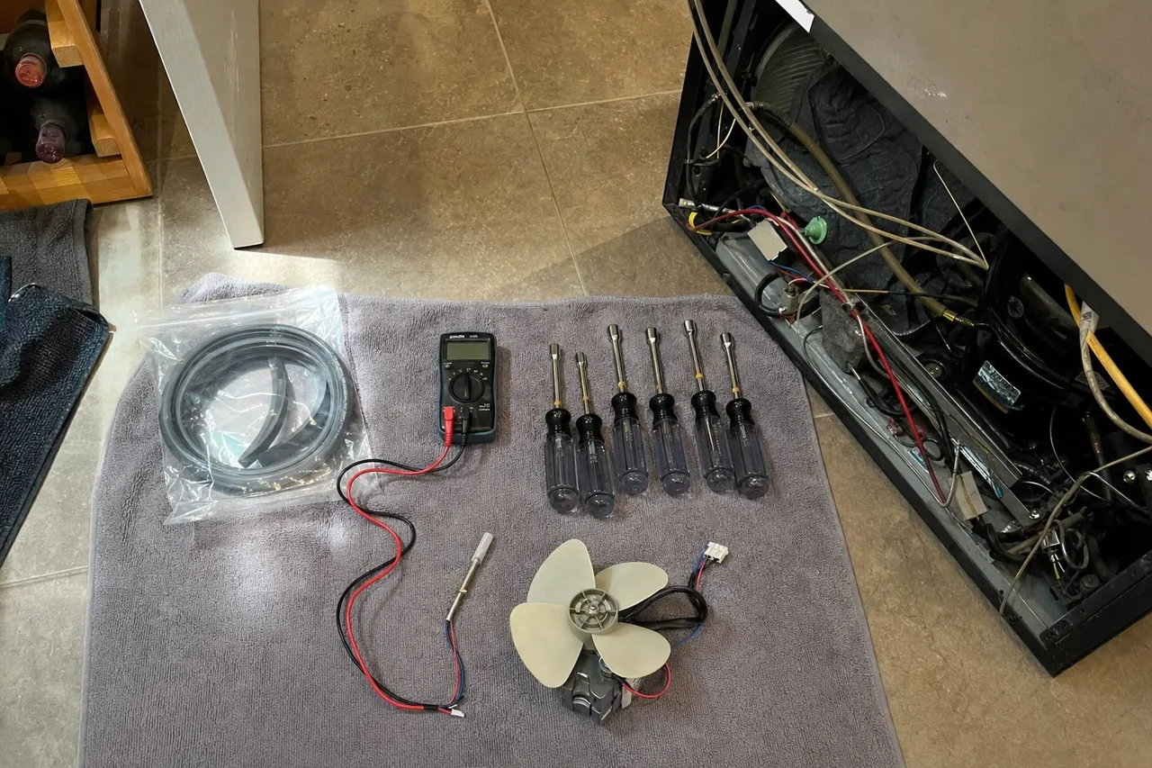 A digital multimeter with its red and black test leads uncoiled on a towel, next to the open machine compartment of a refrigerator and its bundled wiring loom.