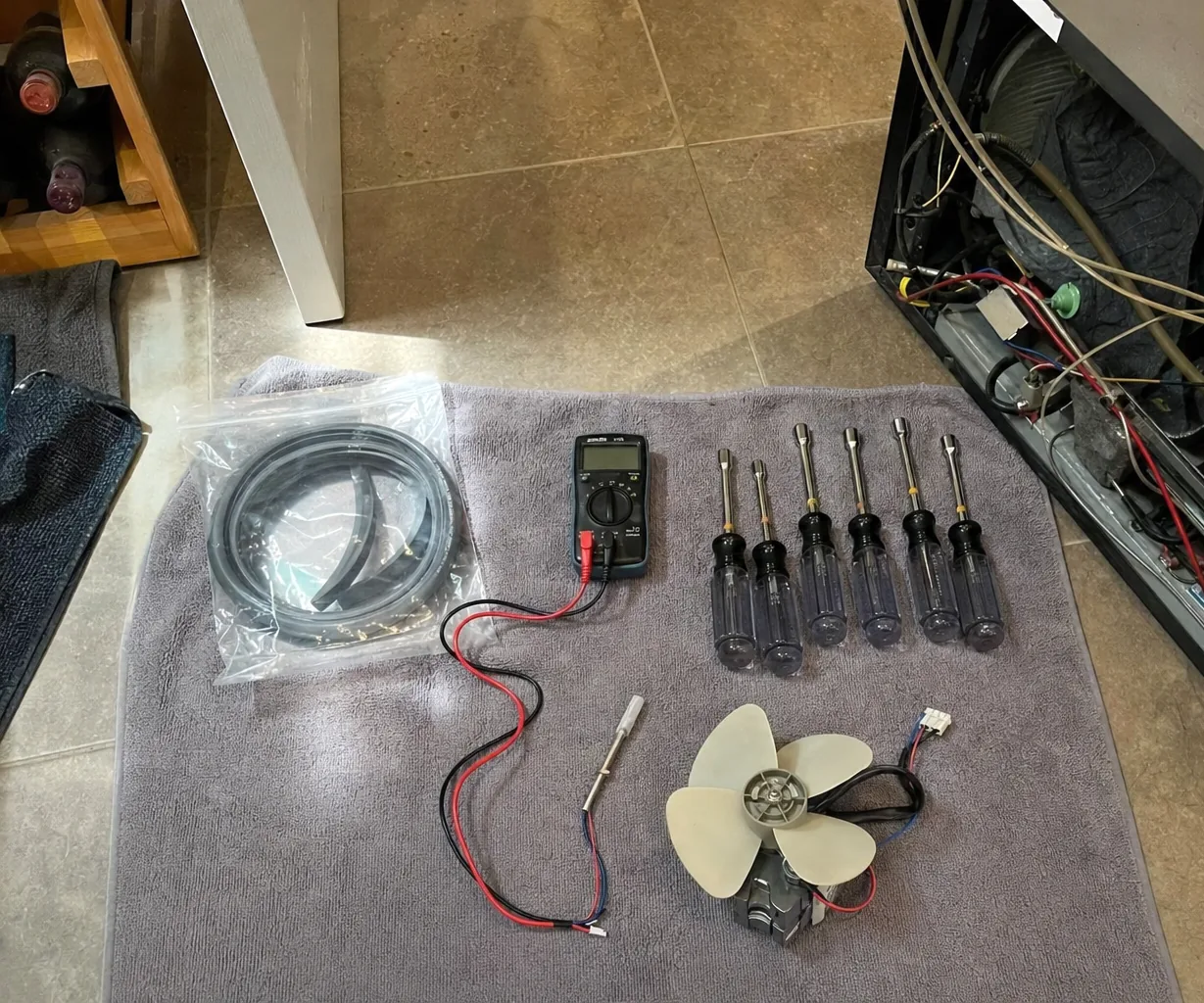 A replacement refrigerator door gasket coiled inside a clear parts bag, resting on a folded towel next to a multimeter, with a wine rack at the edge of the kitchen floor.