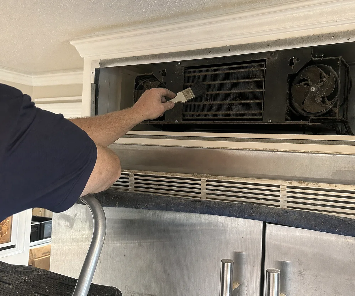 A technician’s bare forearm reaching into the condenser compartment of a built-in refrigerator with a soft brush, the removed grille and a step stool at the edge of the frame.