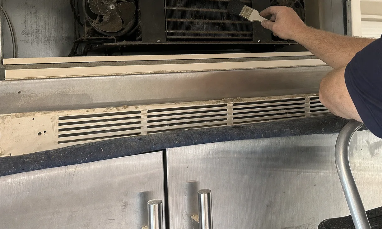 The lower vent slats and stainless door tops of a built-in refrigerator, grey dust caught along the louvres and dulling the metal.
