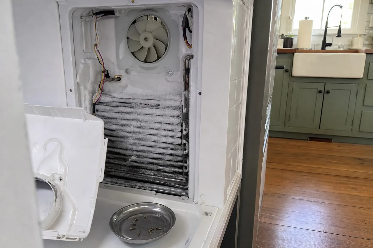 A freezer compartment with its rear cover off, a white fan mounted above frosted evaporator coils, the cover set aside and a steel dish of loose screws on the floor, green cabinets beyond.